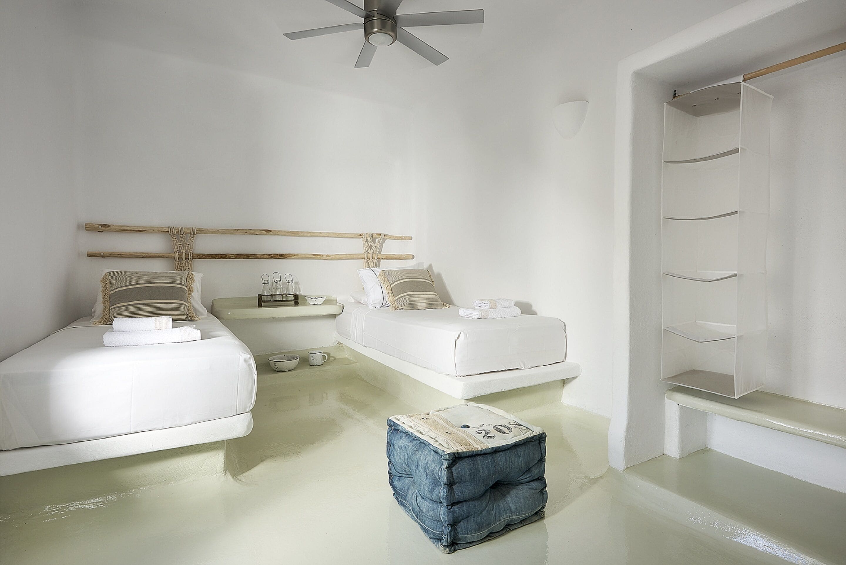 Une chambre à coucher au style minimaliste, avec un lit blanc, une petite table de chevet, un porte-vêtements en bois suspendu et une armoire blanche avec des étagères.