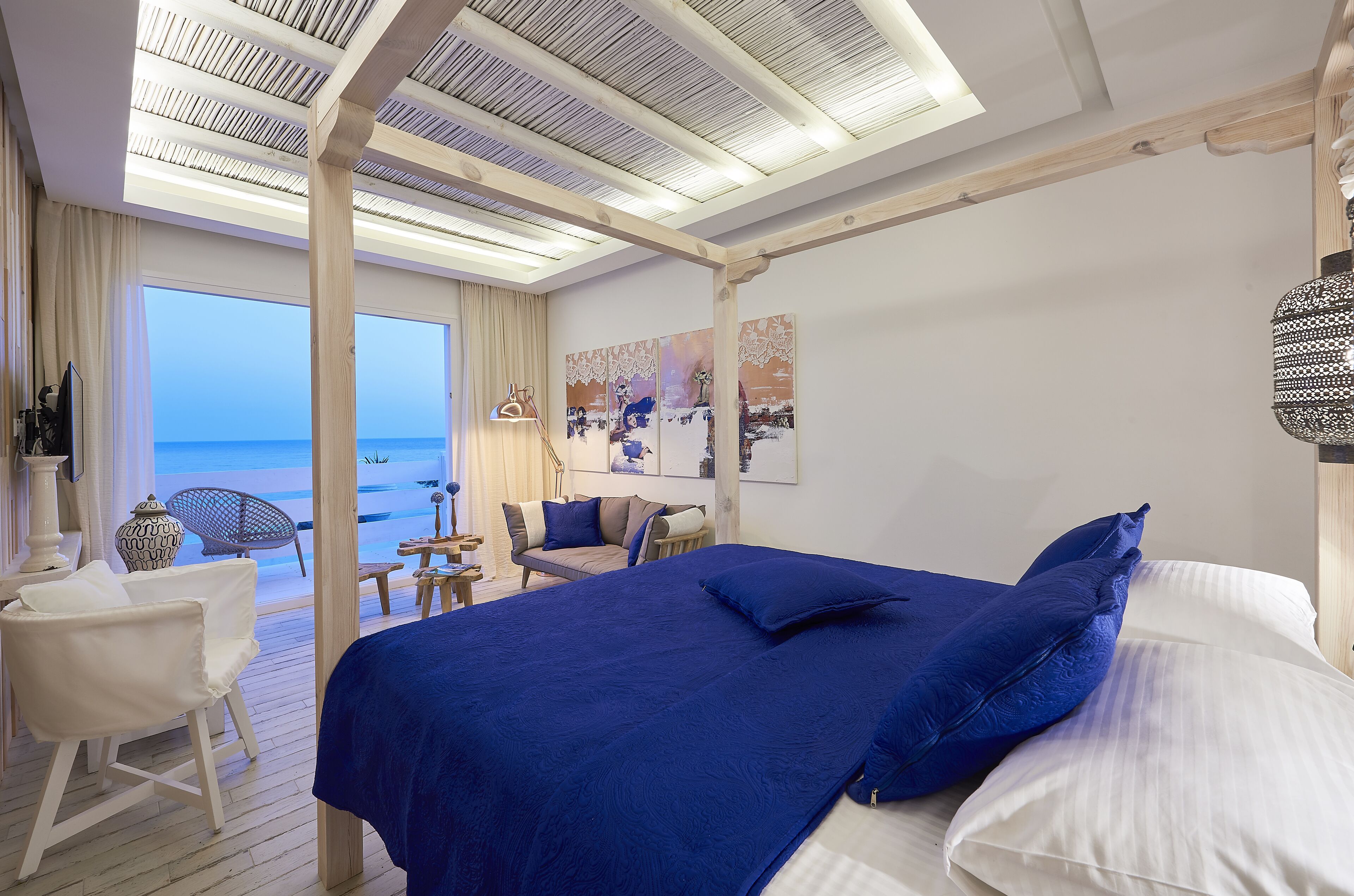 A bedroom with a large bed, a view of the sea, a wooden ceiling, and a seating area with a chair and table.