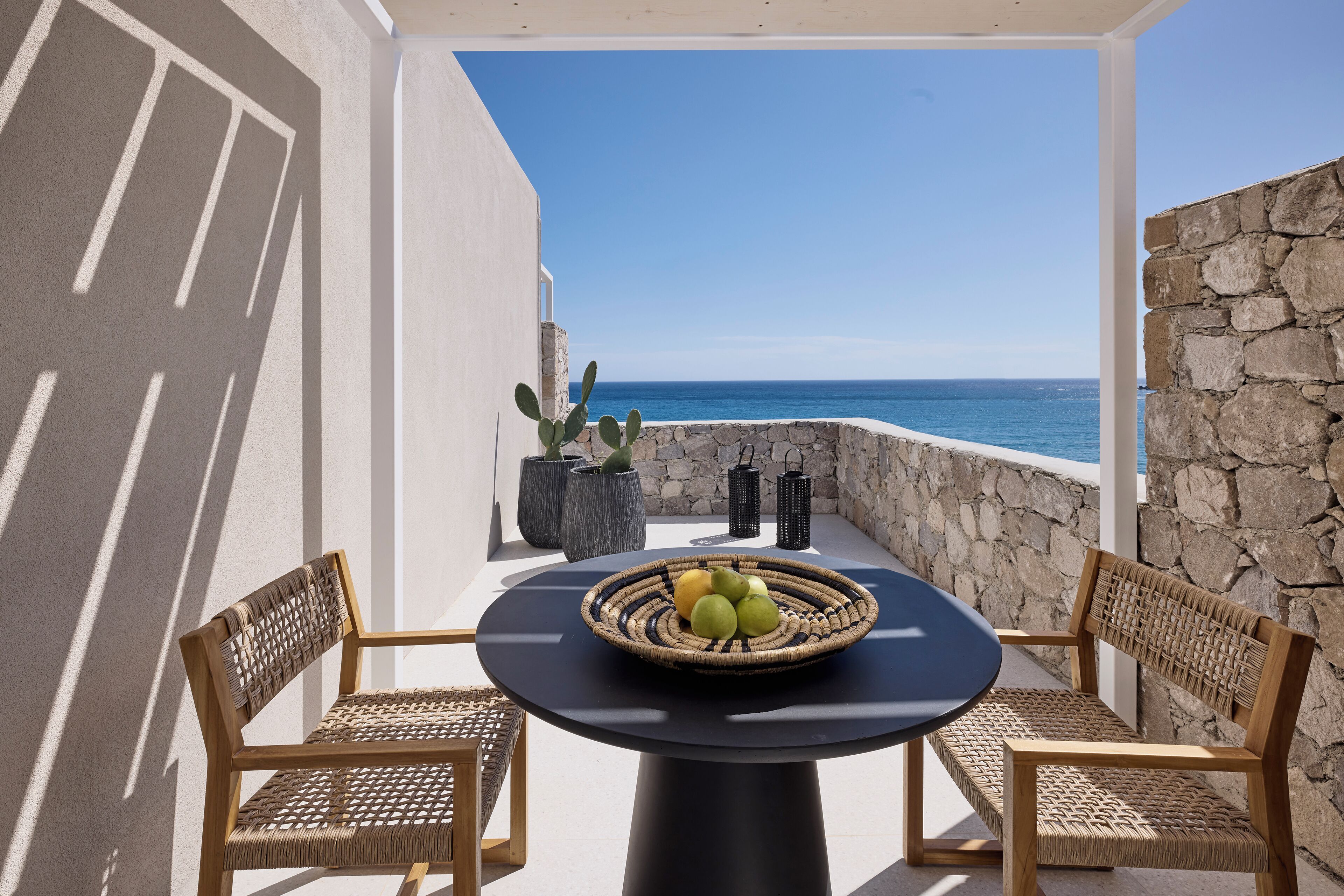 A balcony with wicker chairs, a round table with a woven basket of fruit, and a stone wall overlooking the sea.
