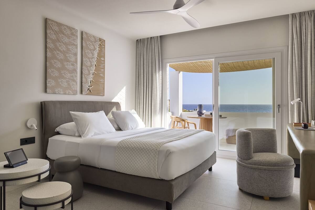 A modern hotel room with a large bed, a desk, a chair, and a balcony with a view of the sea.