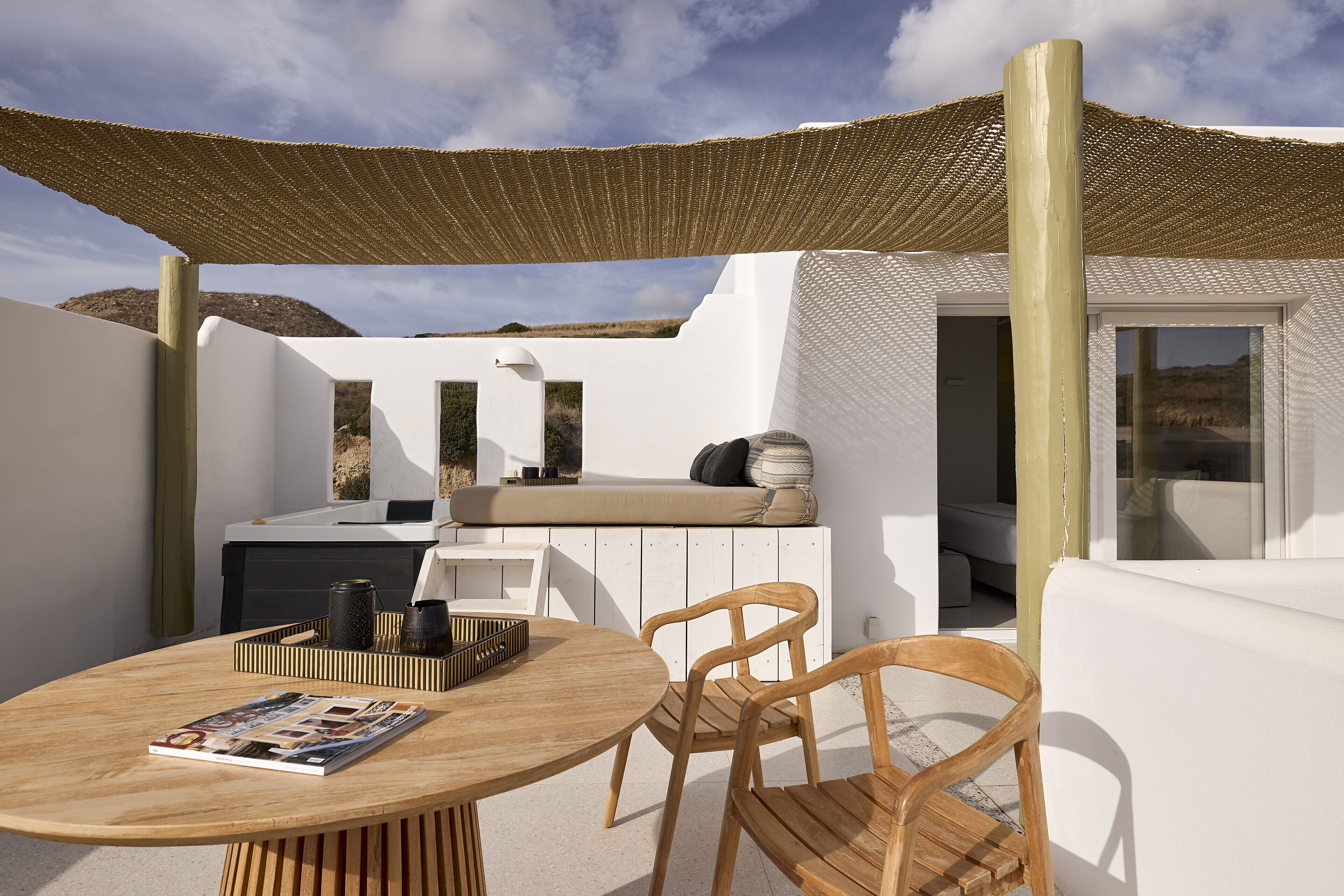 Zona d'estar exterior moderna amb taula rodona de fusta, dues cadires de fusta, una palesa teixida i una paret blanca amb relleu.