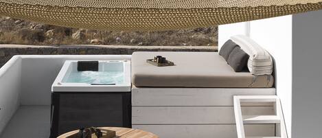 A sunlit outdoor seating area with a hot tub, wooden chairs, and a table with magazines and a tray of drinks.