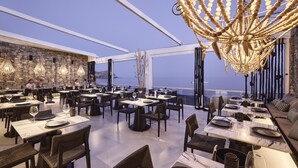 A modern restaurant with a high ceiling, stone wall, and a view of the sea. Tables are set with wine glasses and plates. - Artemis Seaside Resort (Milos)