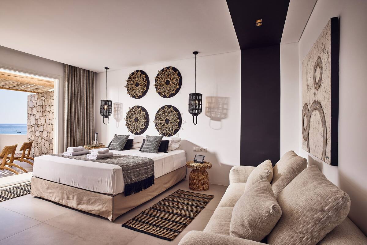 A modern bedroom with a large bed, a sitting area with a sofa, and a view of the sea through a large window.