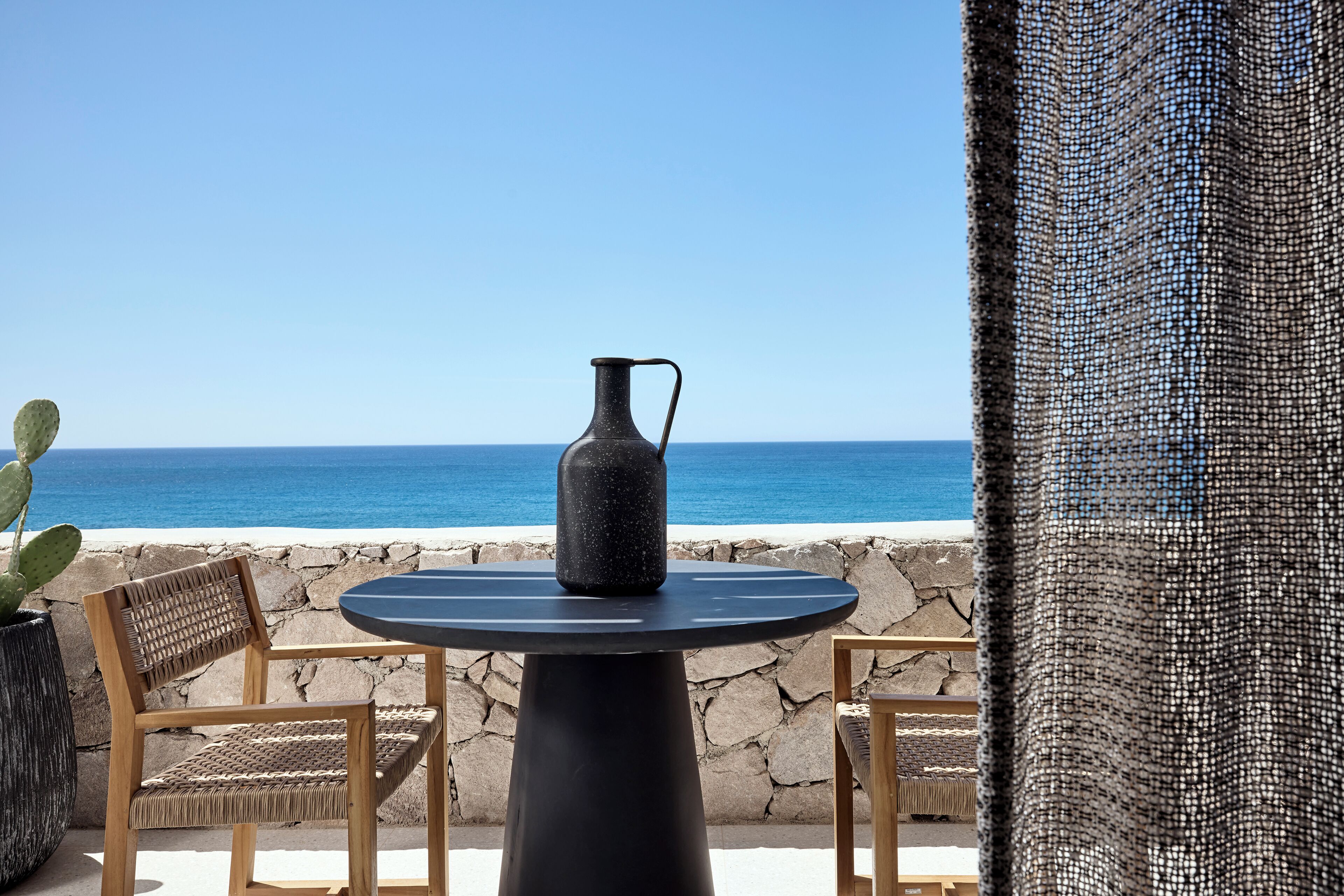 A table with a pitcher, two wicker chairs, and a view of the ocean.