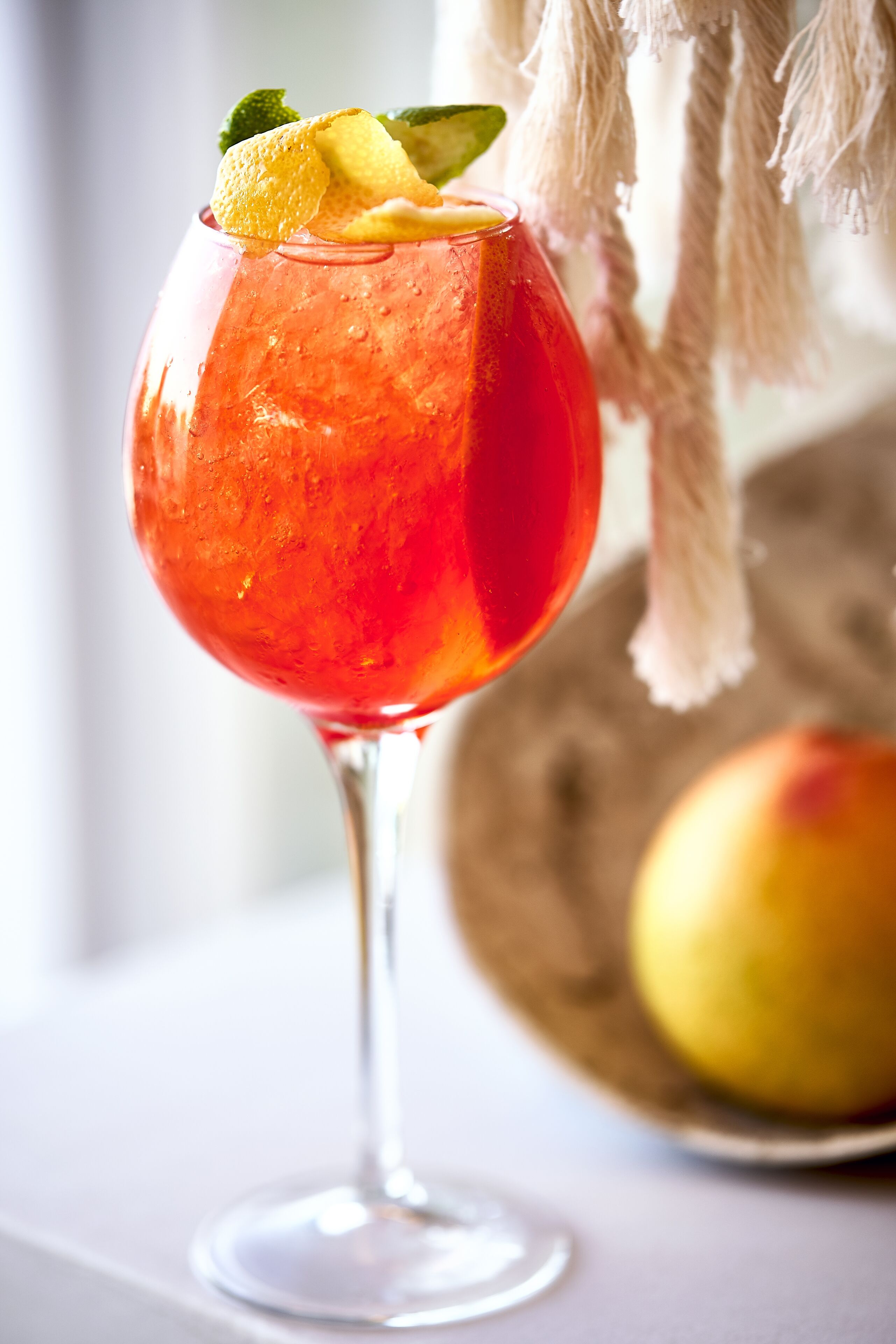 A cocktail in a stemmed glass with a lime wedge garnish, set against a blurred background with a fruit and a hanging decoration.
