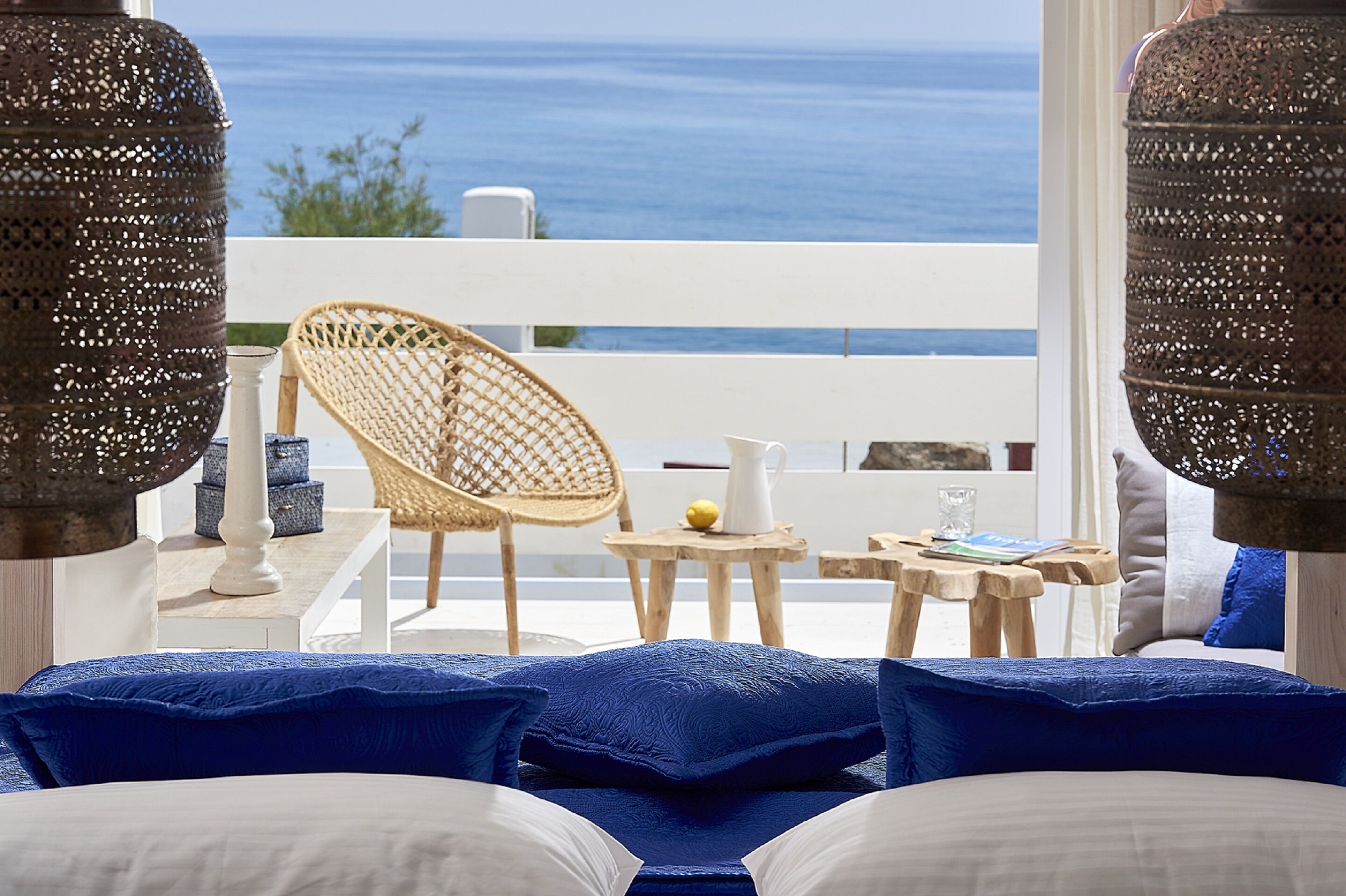 A bedroom with a view of the sea, a wicker chair, a small table with a pitcher, and a white railing.