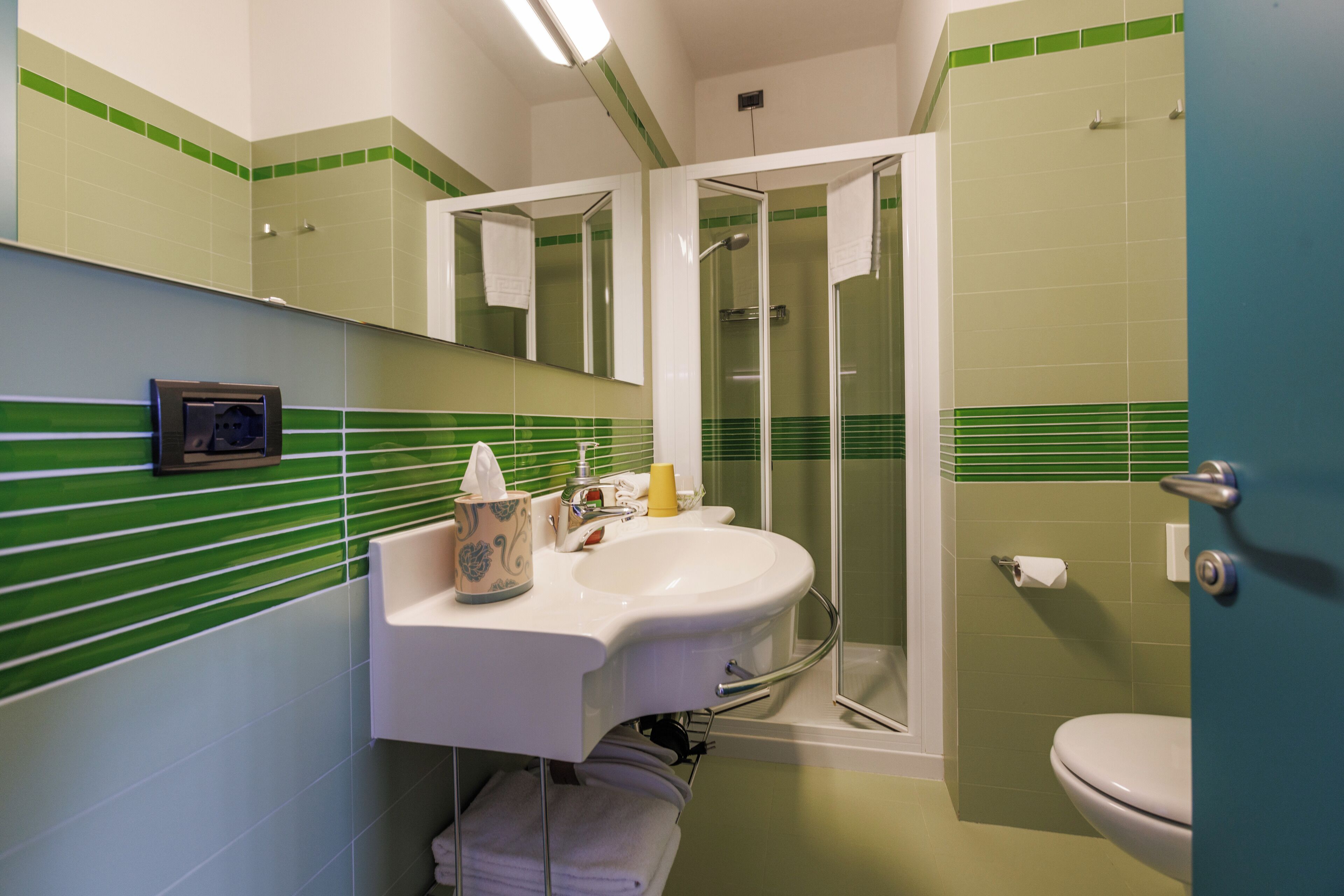 Een badkamer met een wit wastafelmeubel, tegels met groene strepen, een douche met glazen wand en een toilet.