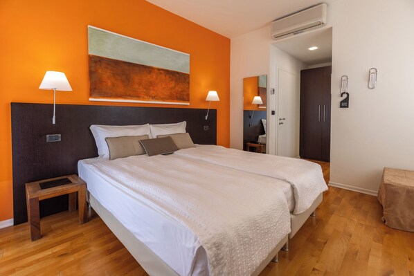 A hotel room with a large bed, two bedside lamps, a small wooden nightstand, a painting on the wall, and an air conditioning unit.