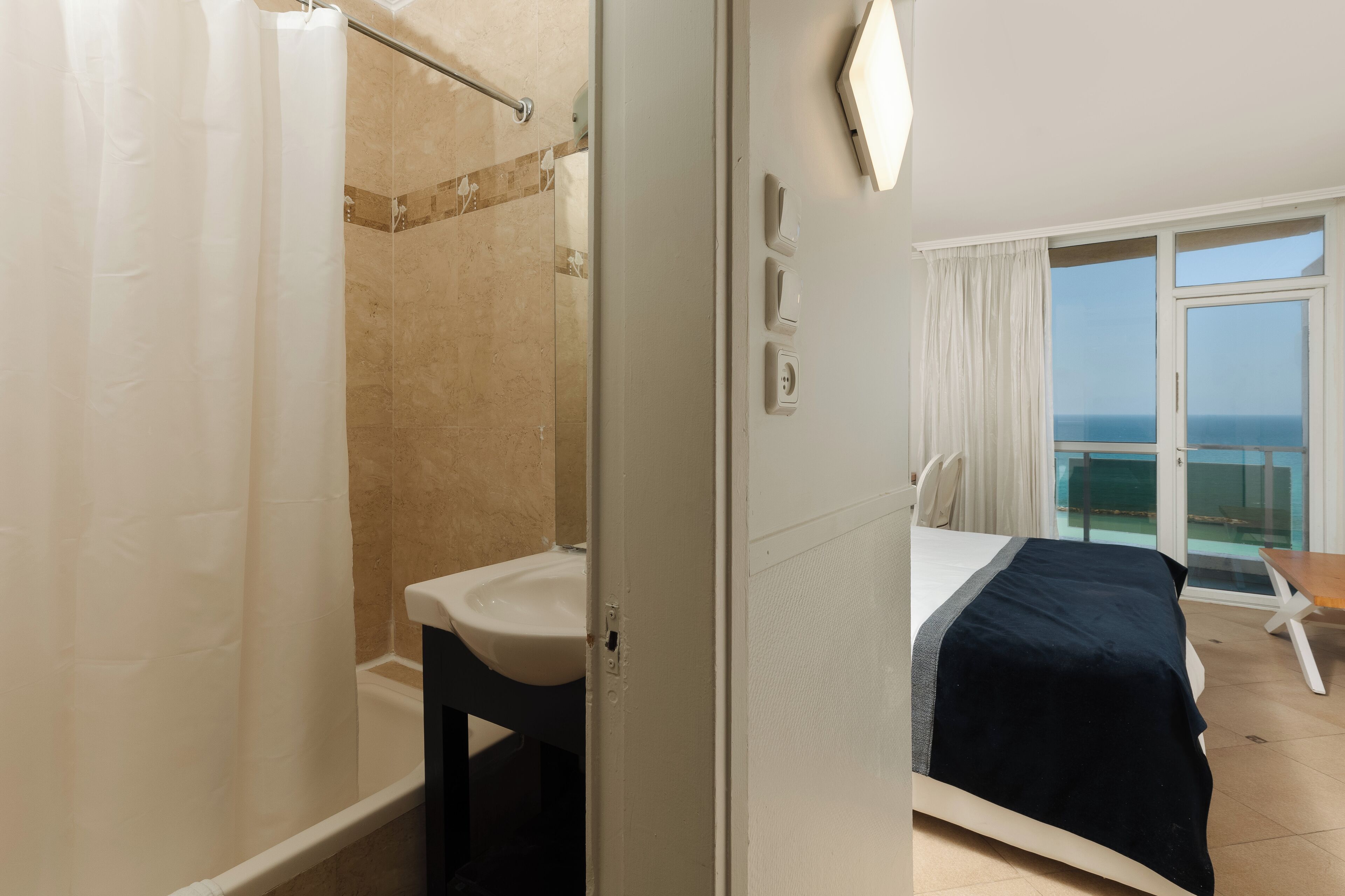 A hotel room with a bathroom, a bed, a balcony with a view of the sea, and a window with curtains.