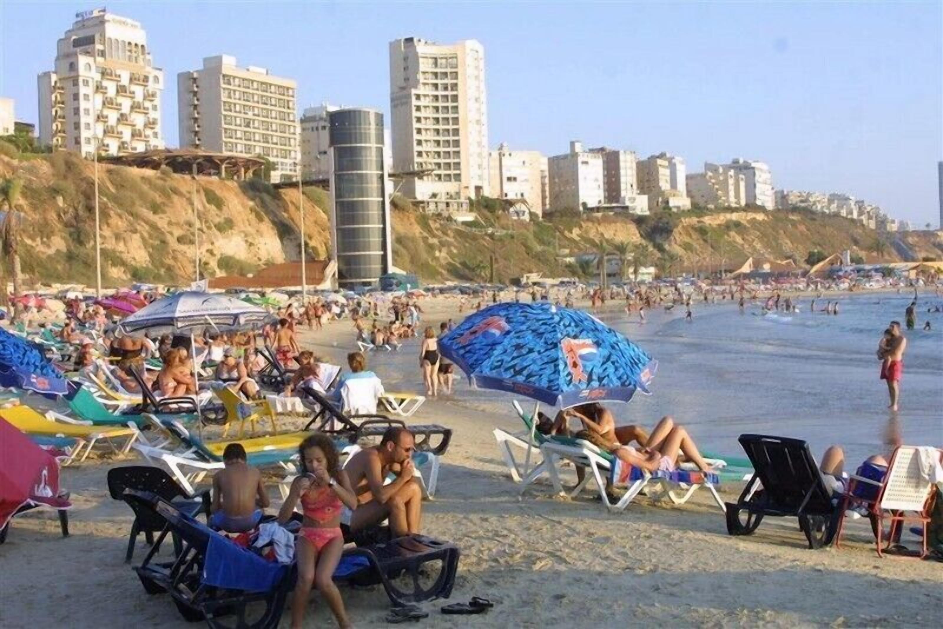 A crowded beach with numerous sun loungers and umbrellas, a sandy shore, and buildings in the background.
