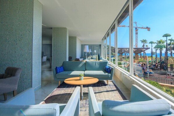 Un espacio lounge moderno con un sofá, sillones, una mesa de centro y vistas a una obra en construcción y palmeras.