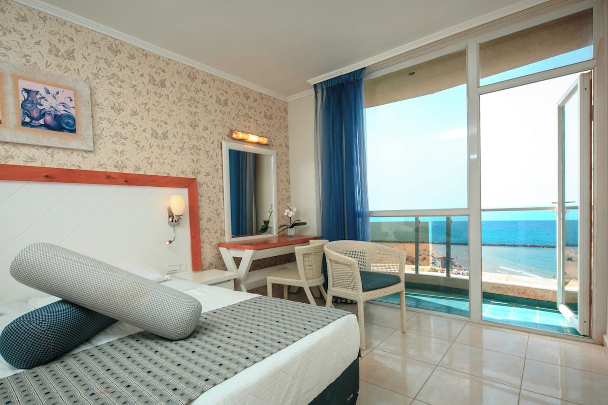 A hotel room with a bed, a desk, a chair, a large window with a view of the sea, and a balcony.