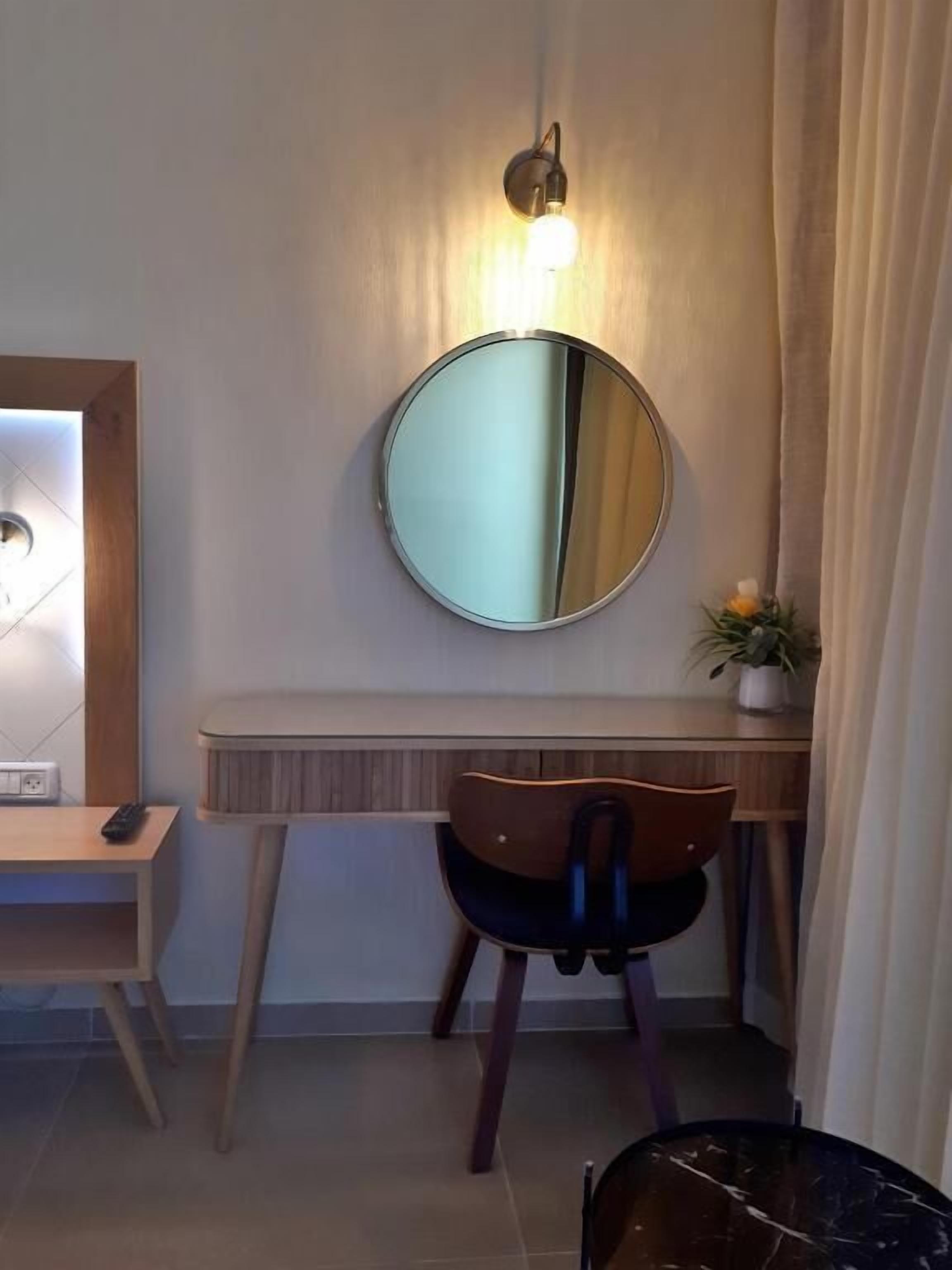 A modern room with a round mirror, a desk, a chair, and a wall-mounted light fixture.