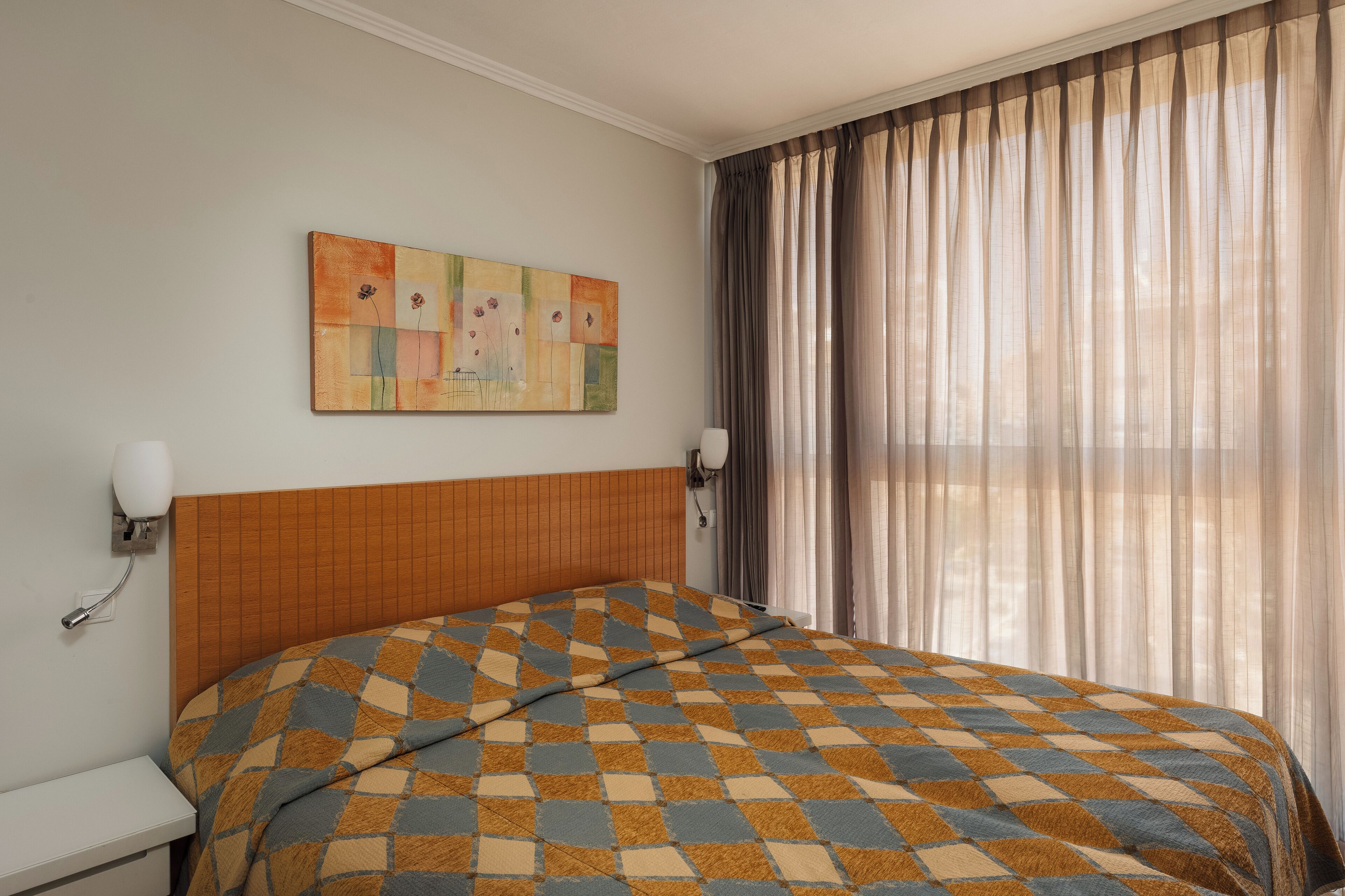 A hotel room with a checkered bedspread, a wall-mounted painting, a bedside lamp, and a window with sheer curtains.