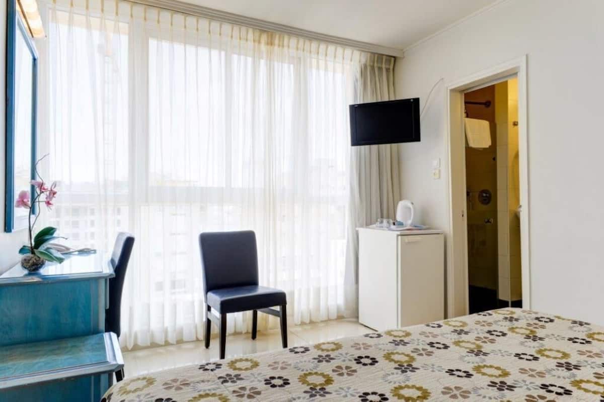 A hotel room with a bed, a desk with a chair, a television mounted on the wall, and a bathroom visible through an open door.