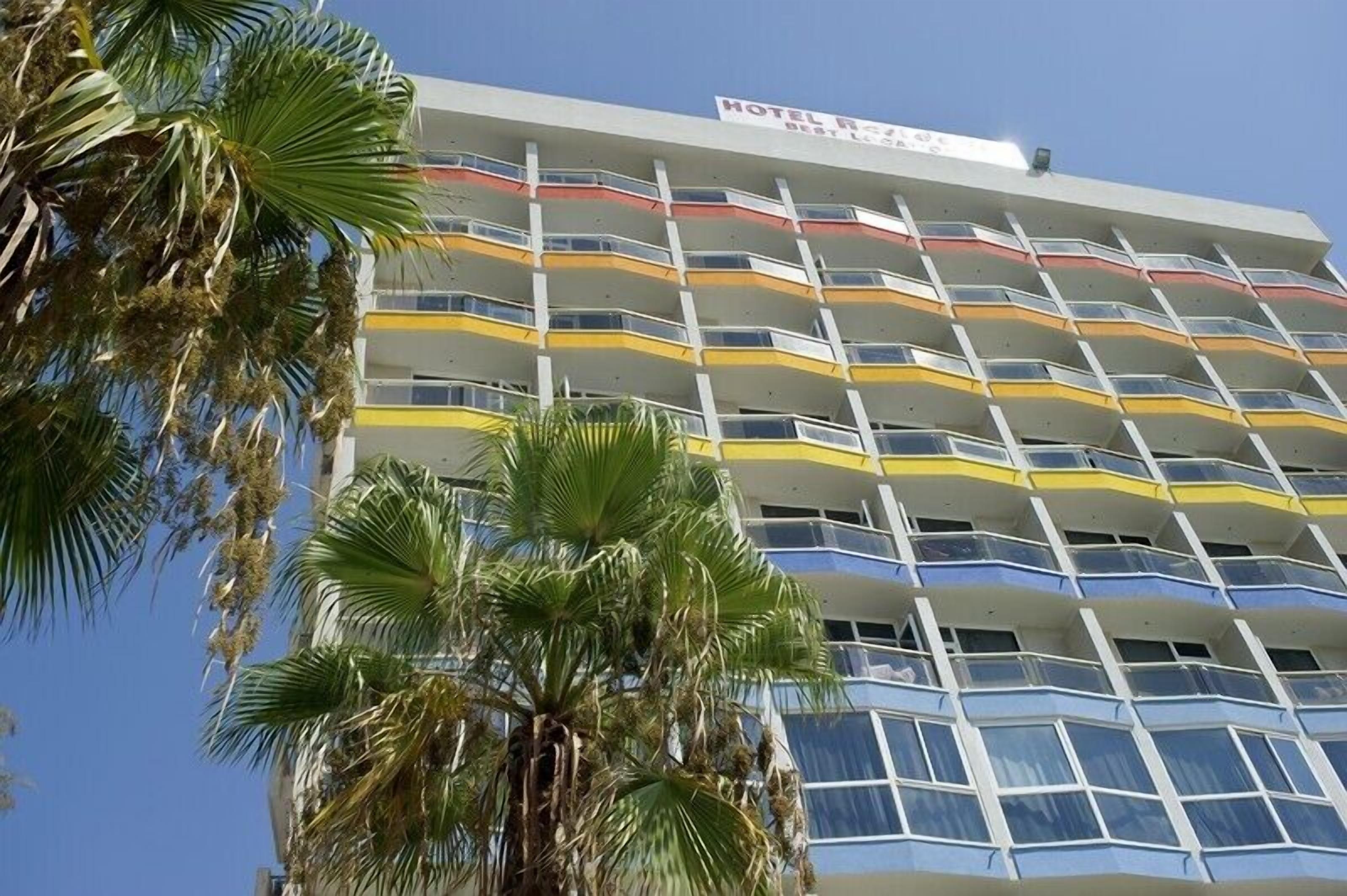 A multi-story hotel with balconies, palm trees in the foreground, and a clear blue sky.