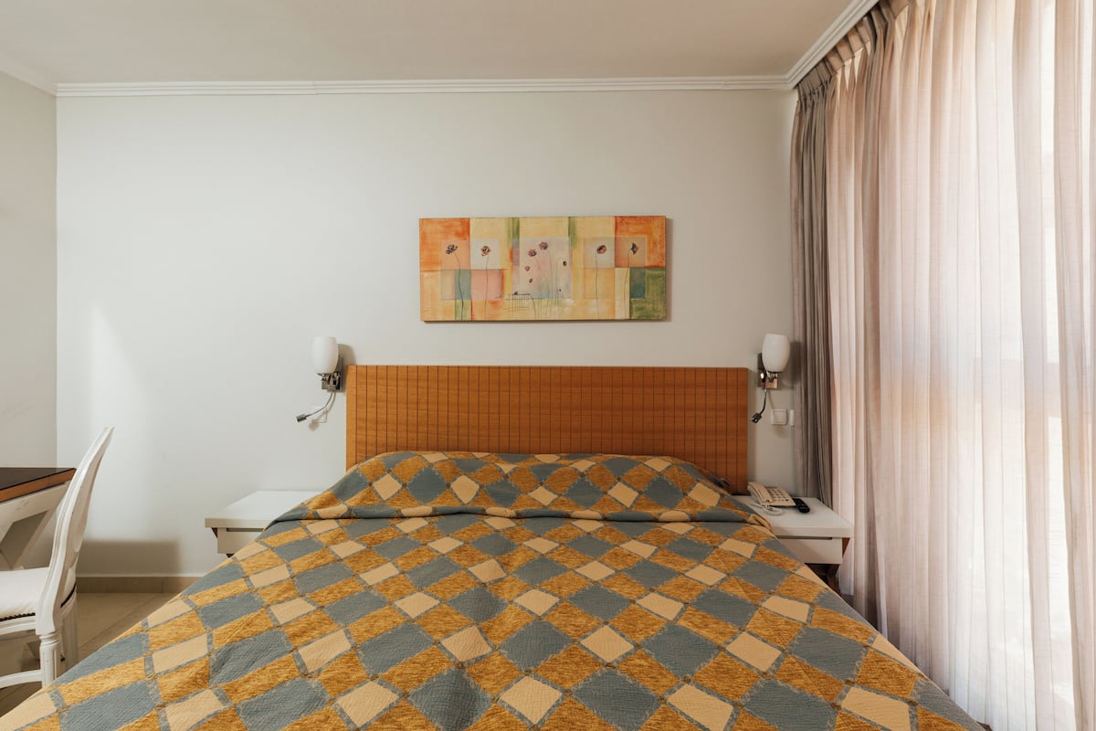 A hotel room with a bed, a desk, a chair, a telephone, and a painting on the wall.
