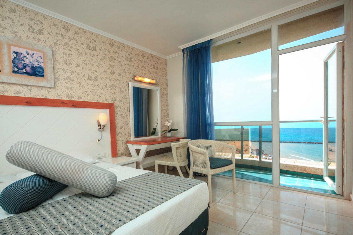 A hotel room with a bed, a desk, a chair, a large window with a view of the sea, and a balcony.
