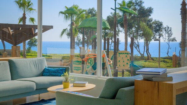 Una habitación moderna con un ventanal grande, un sofá, una mesita con un libro y vistas al mar y palmeras.