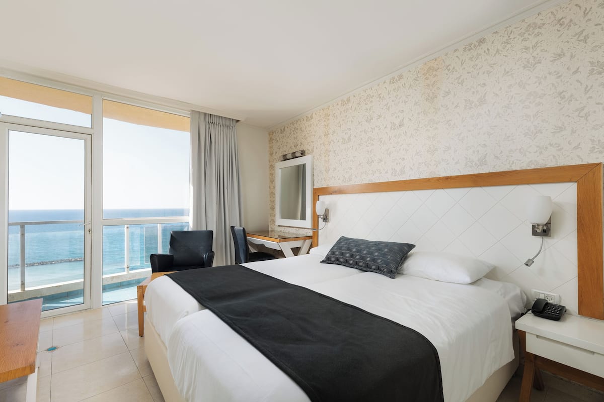 A hotel room with a large bed, a desk with a chair, a small table, a nightstand with a phone, and a view of the ocean through a sliding door.