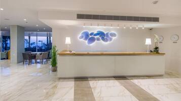 Un área de recepción moderna en un hotel, con suelo de mármol, mostrador de recepción y una zona de estar con sillas y una mesa.