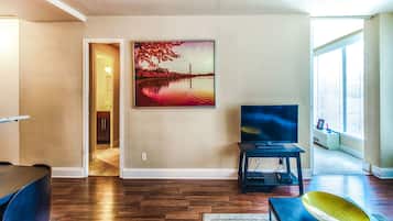 A living room with hardwood flooring, a flat-screen TV on a stand, a painting of a sunset over a body of water, and a view of a cityscape through a large window.