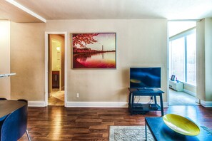 A living room with hardwood flooring, a flat-screen TV on a stand, a painting of a sunset over a body of water, and a view of a cityscape through a large window.