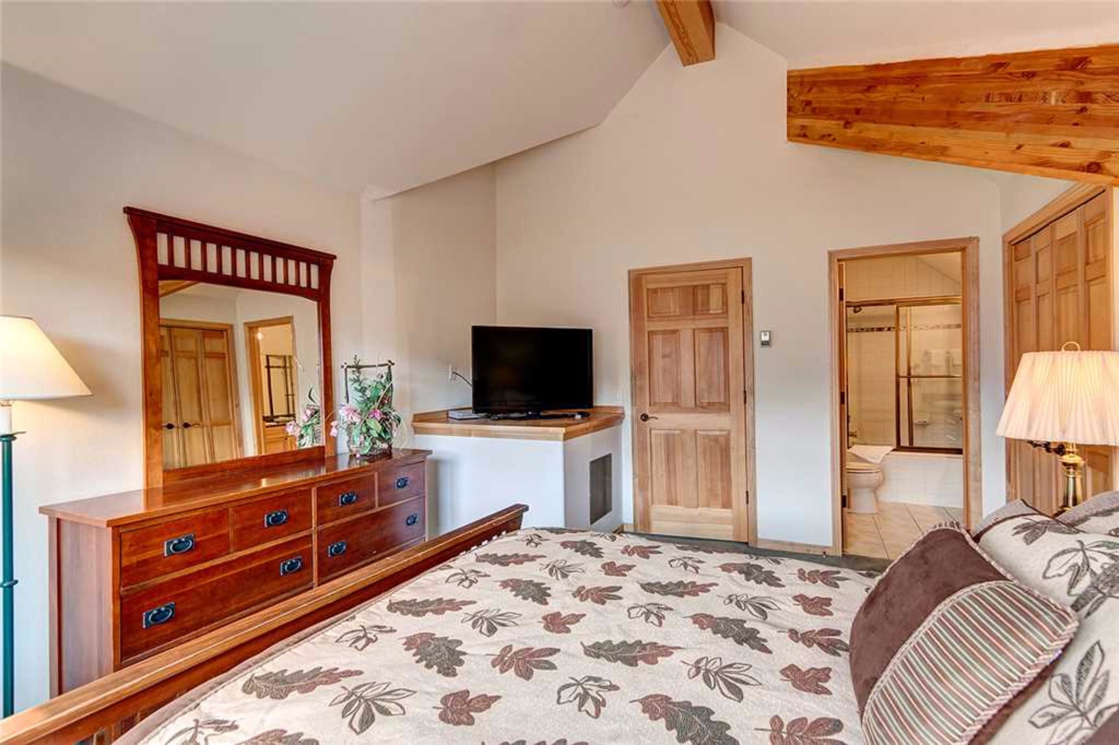 A bedroom with a wooden dresser, a television, a bed with floral bedding, and a wooden ceiling.