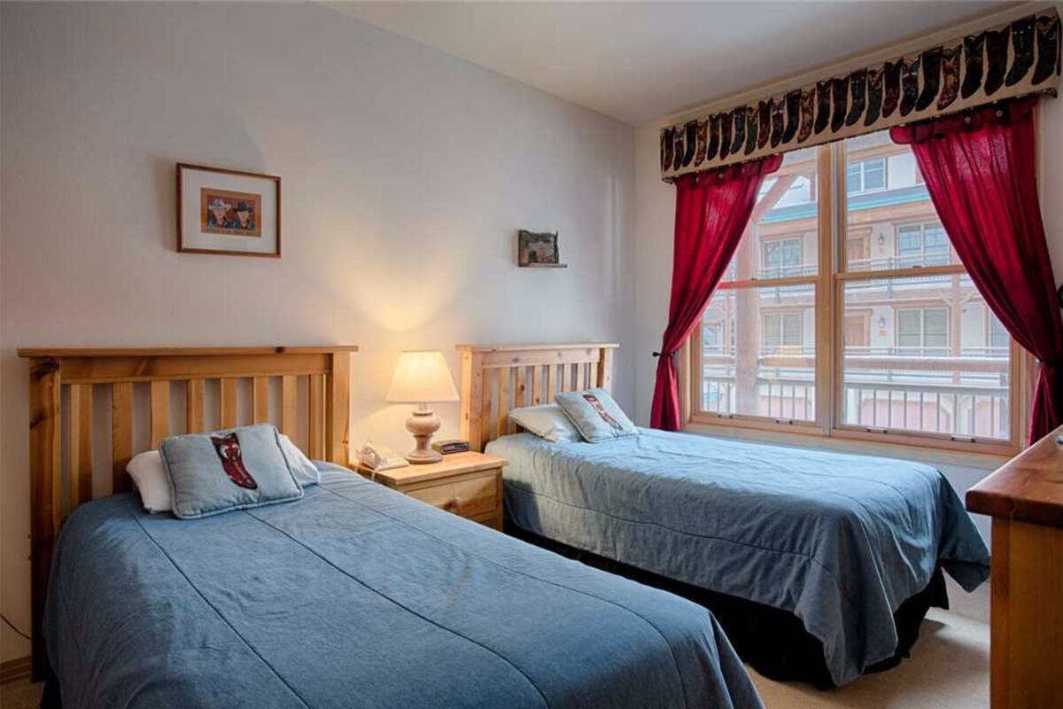 Ein Zimmer mit zwei Betten, einem hölzernen Kopfstück, einem Nachttisch mit Lampe und einem Fenster mit roten Vorhängen.