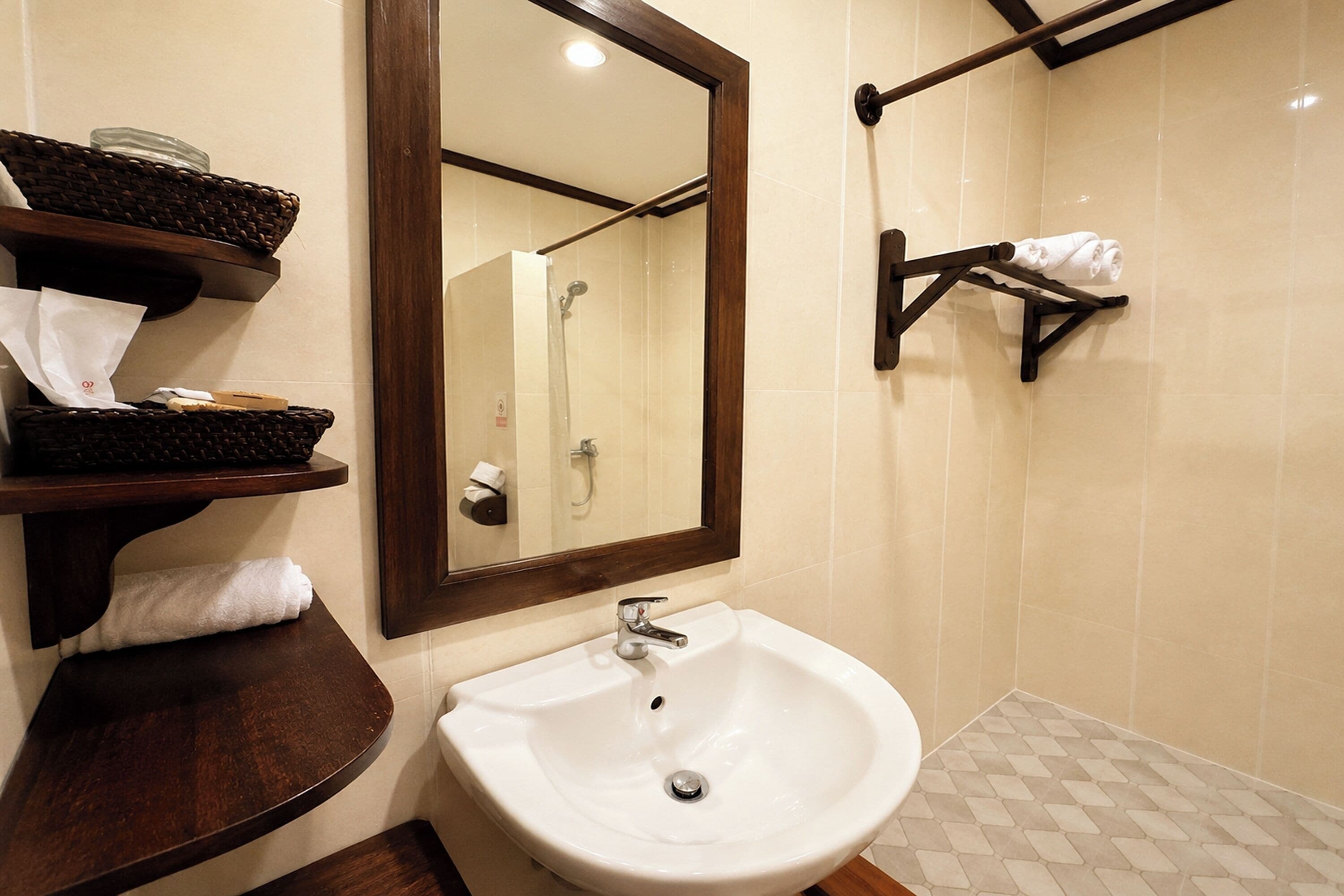 Un baño con un lavamanos blanco, un espejo y una ducha. Hay repisas de madera con toallas y un frasco de vidrio.