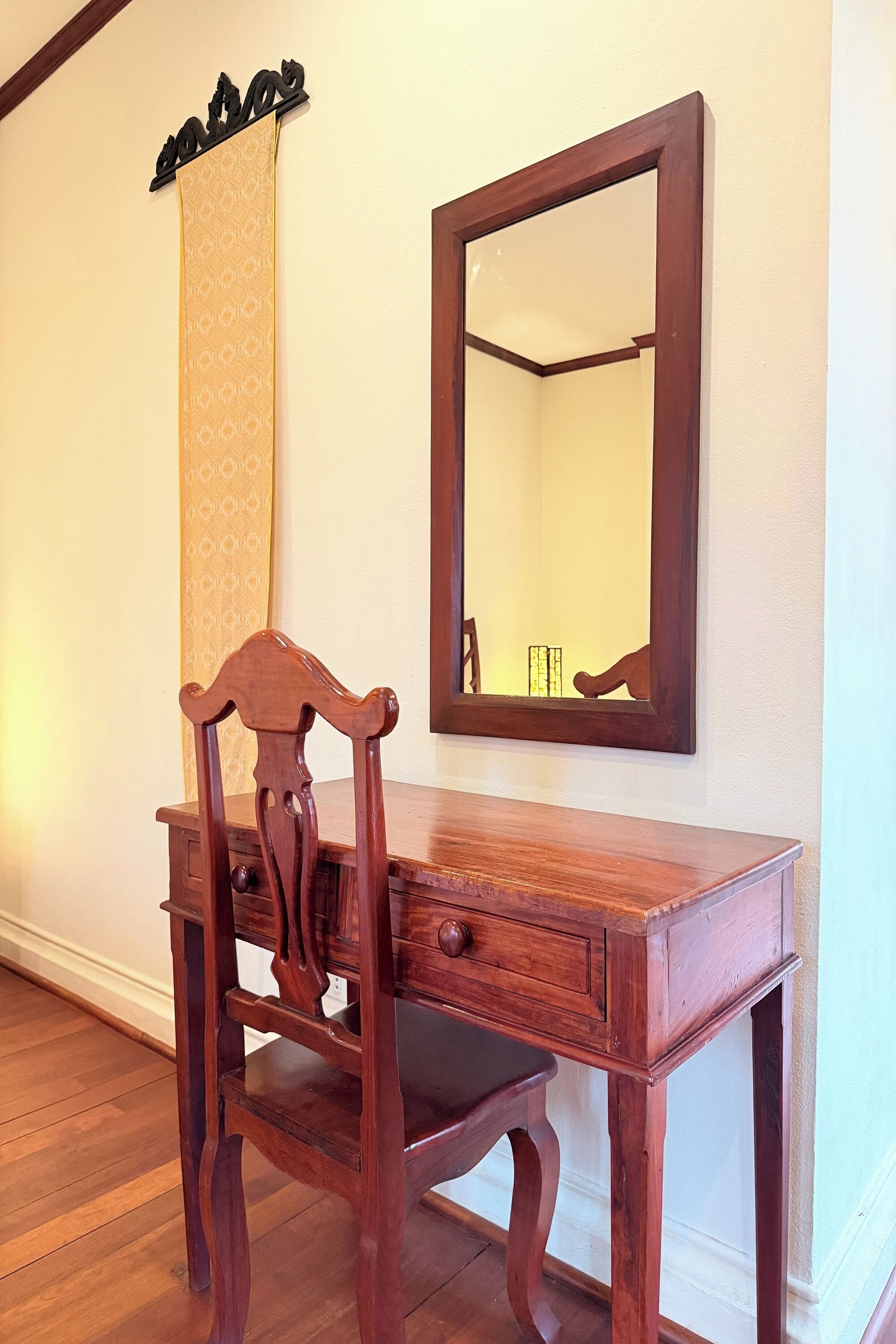Un bureau en bois, avec un miroir et une chaise, dans une pièce dont le sol est recouvert de planches de bois et les murs sont peints en blanc.