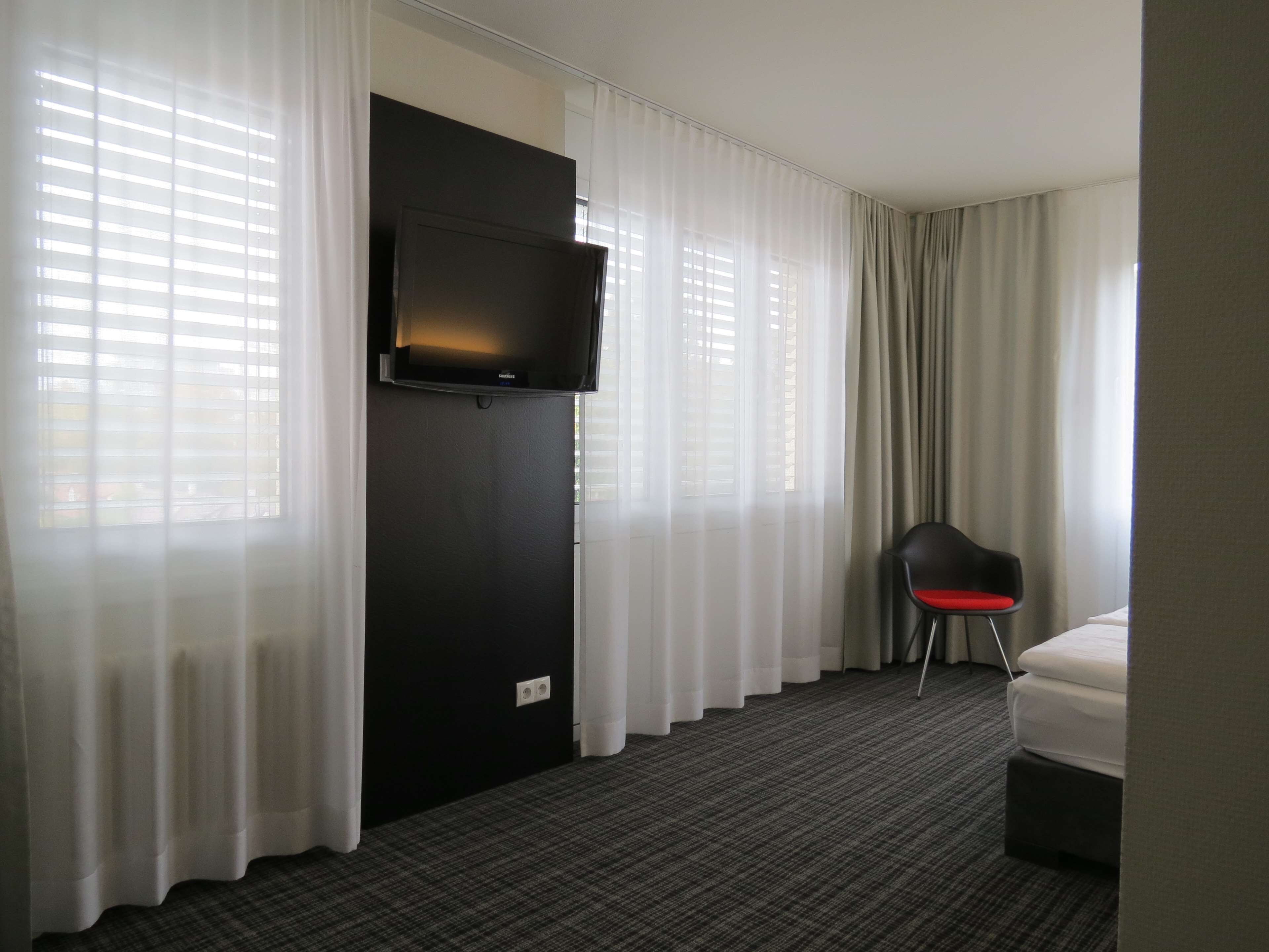 Une chambre d’hôtel avec un téléviseur à écran plat fixé à un mur noir, un lit avec une literie blanche, une chaise noire avec un coussin rouge et des rideaux transparents aux fenêtres.