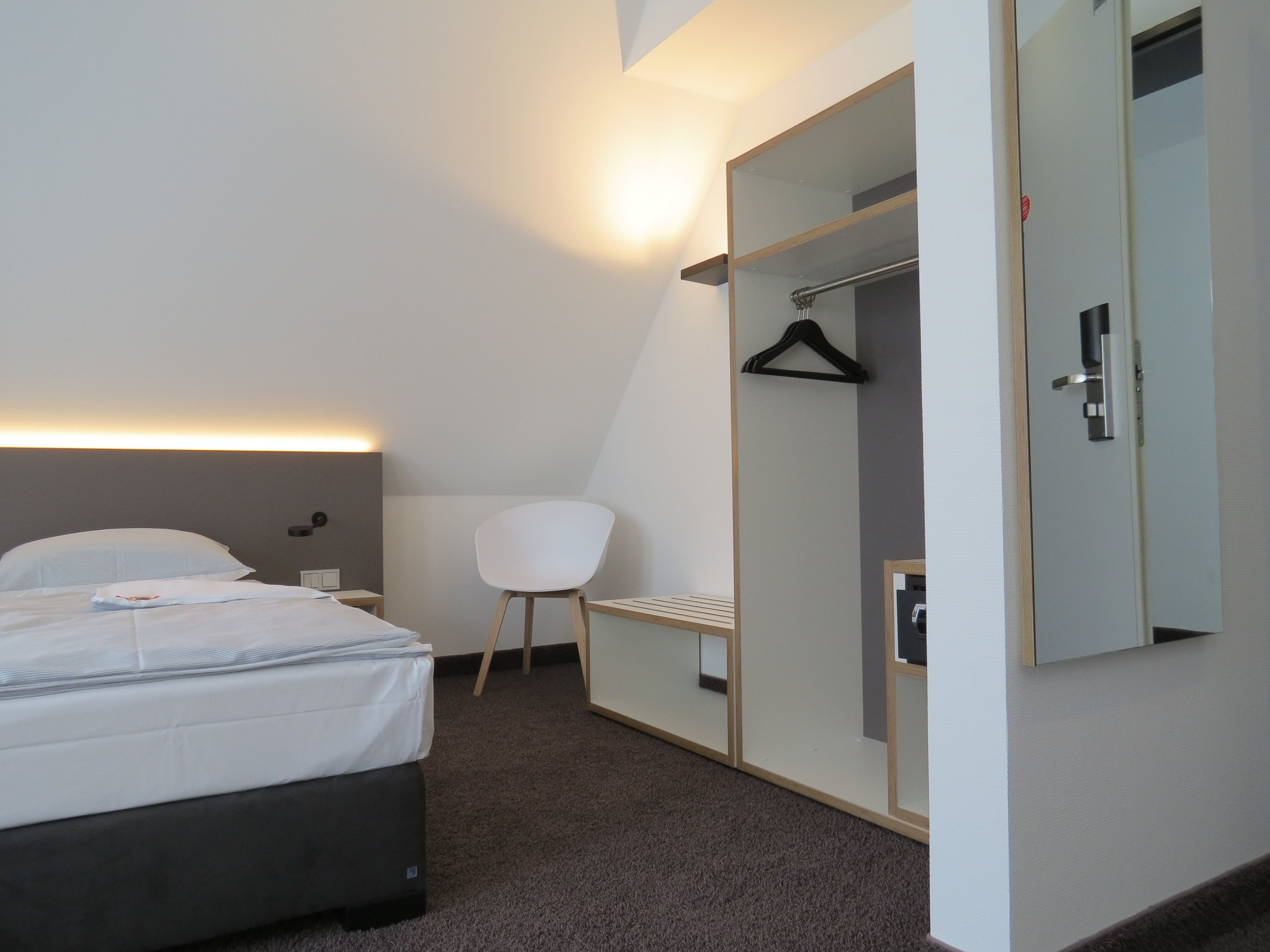 A modern hotel room with a bed, a chair, a wardrobe, and a mirror.