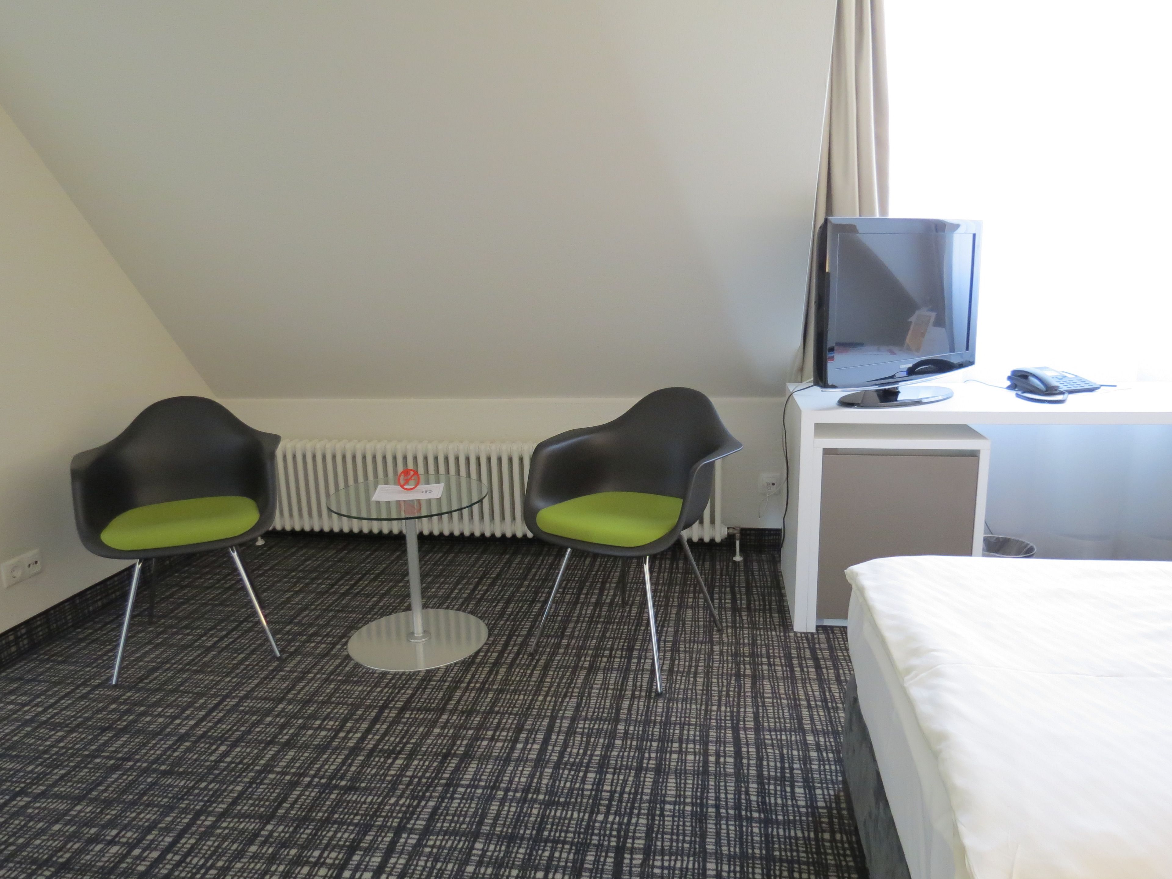 Habitación de hotel con una cama, un escritorio con ordenador y teléfono, dos sillas, una mesita y un radiador.