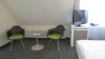 Habitación de hotel con una cama, un escritorio con ordenador y teléfono, dos sillas, una mesita y un radiador.