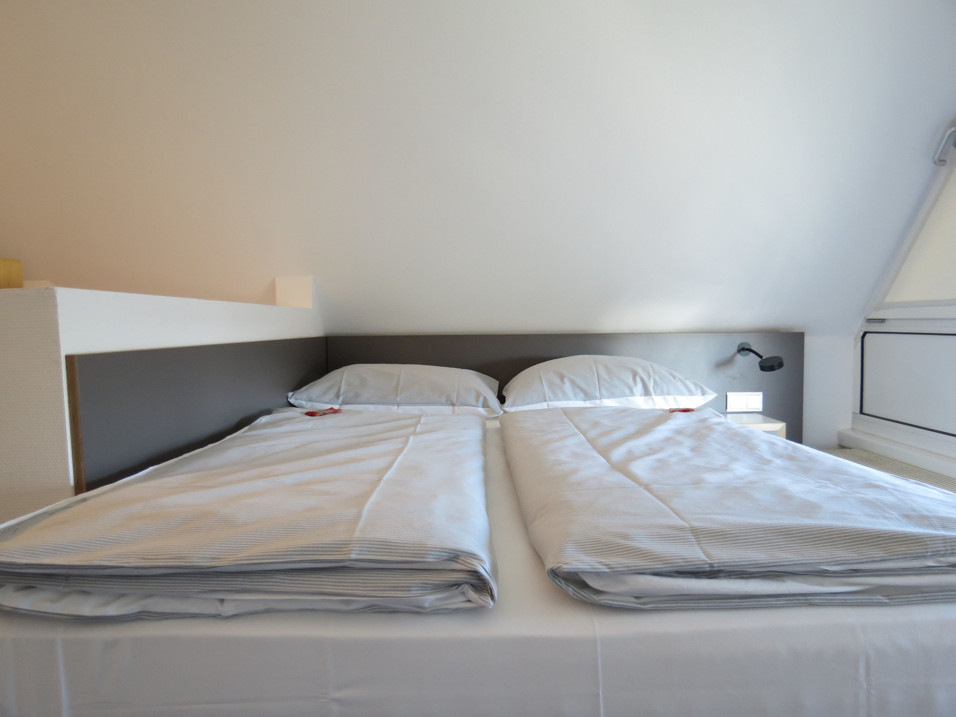 Una cama con dos plazas, un cabecero gris y ropa de cama blanca en una habitación con techo inclinado.