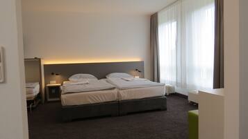 A hotel room with two beds, a bedside table, a nightstand, a window with curtains, and a green ottoman.