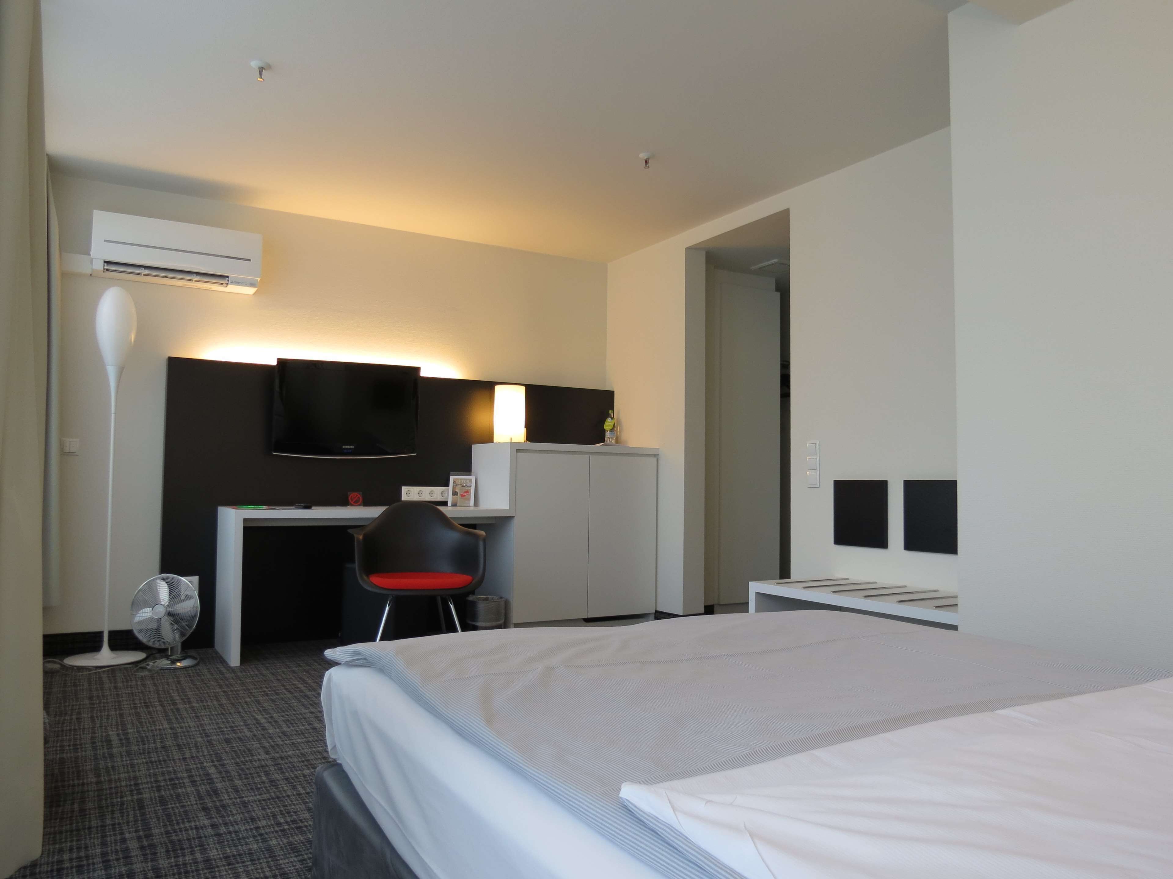 A hotel room with a bed, a desk, a chair, a TV, and an air conditioning unit.