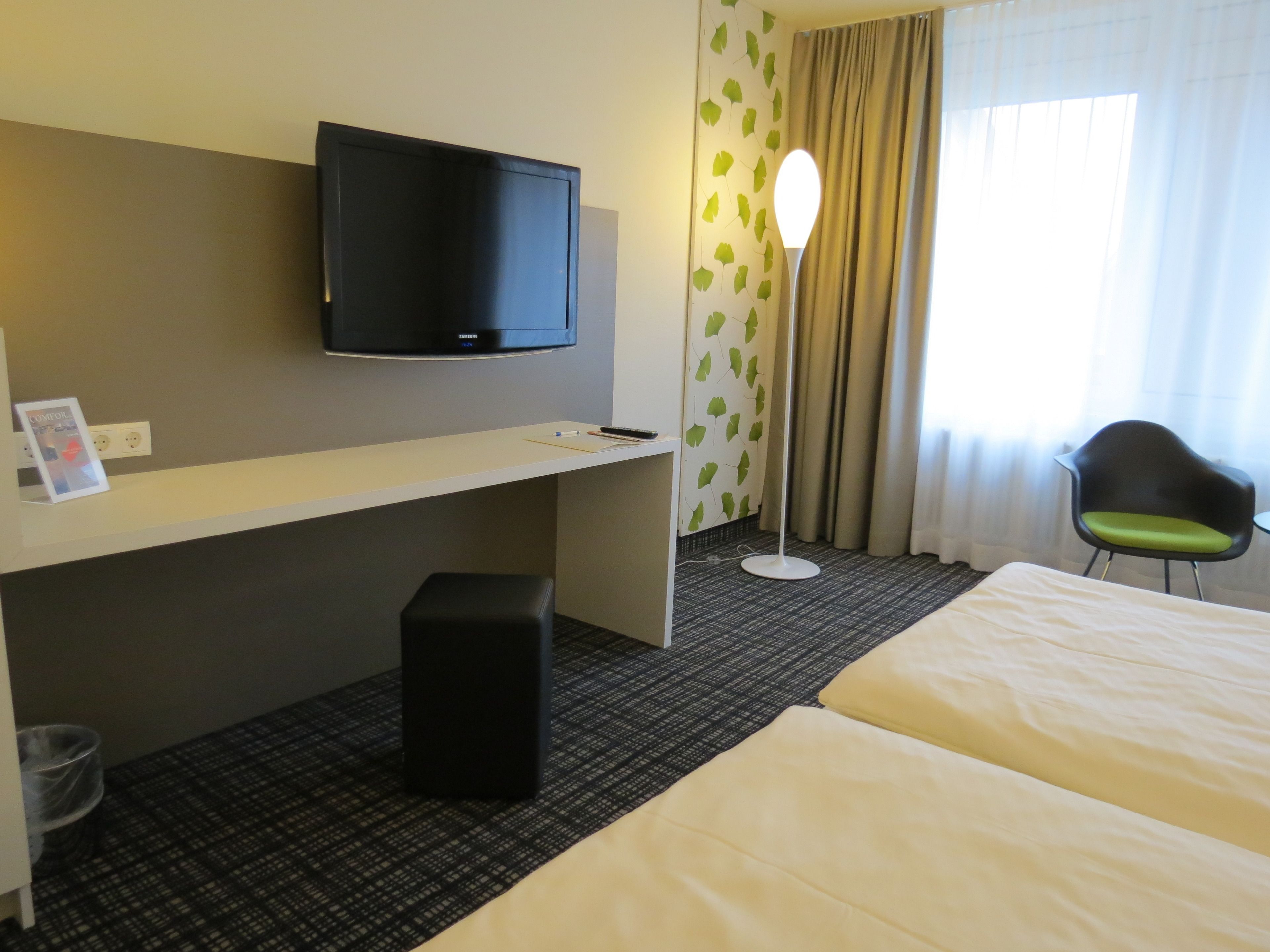 A hotel room with a bed, a desk with a flat-screen TV, a chair, a standing lamp, and a window with curtains.