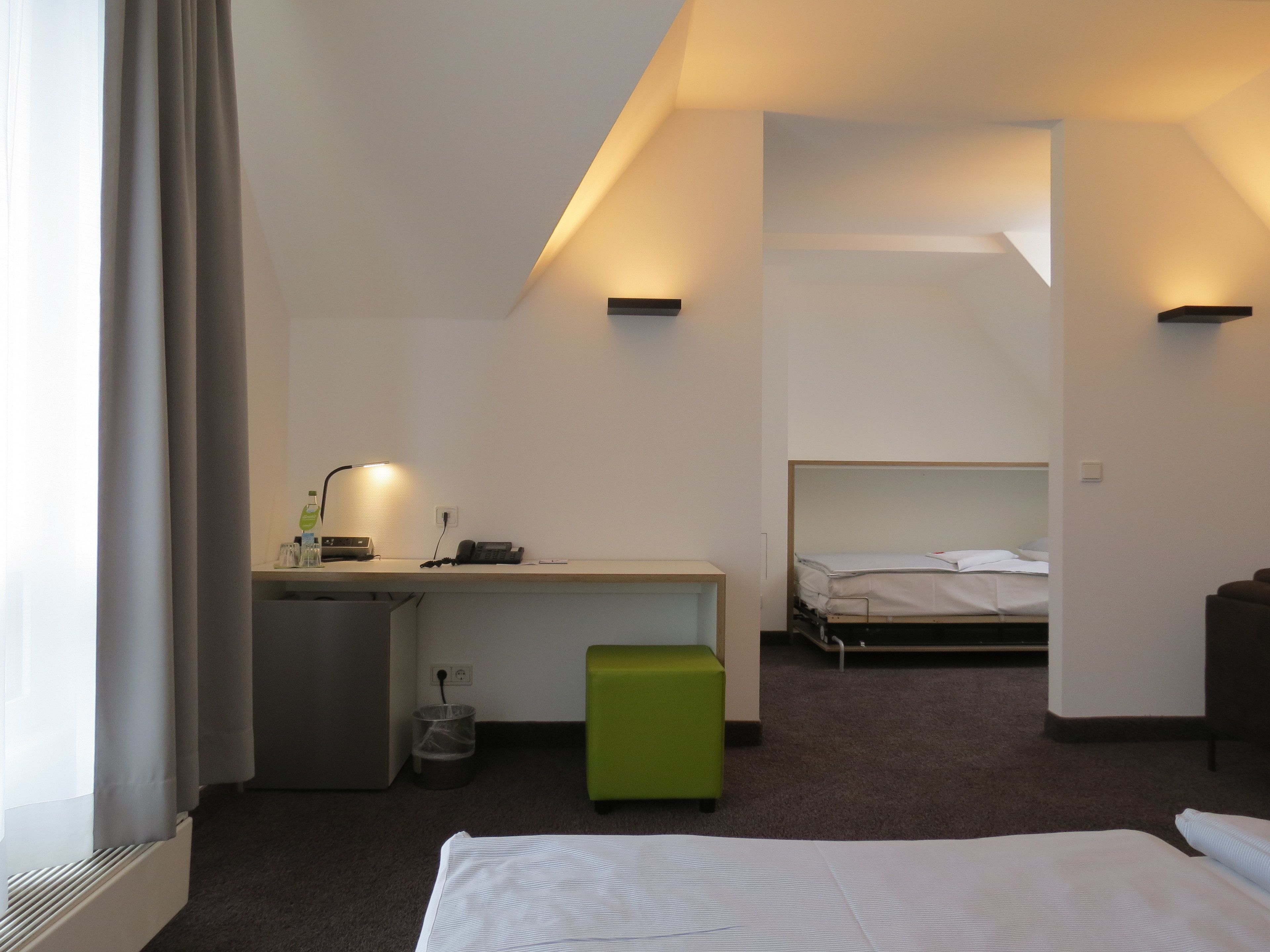 Una habitación de hotel con escritorio, un otoman verde, una cama y una pequeña zona de kitchenette.