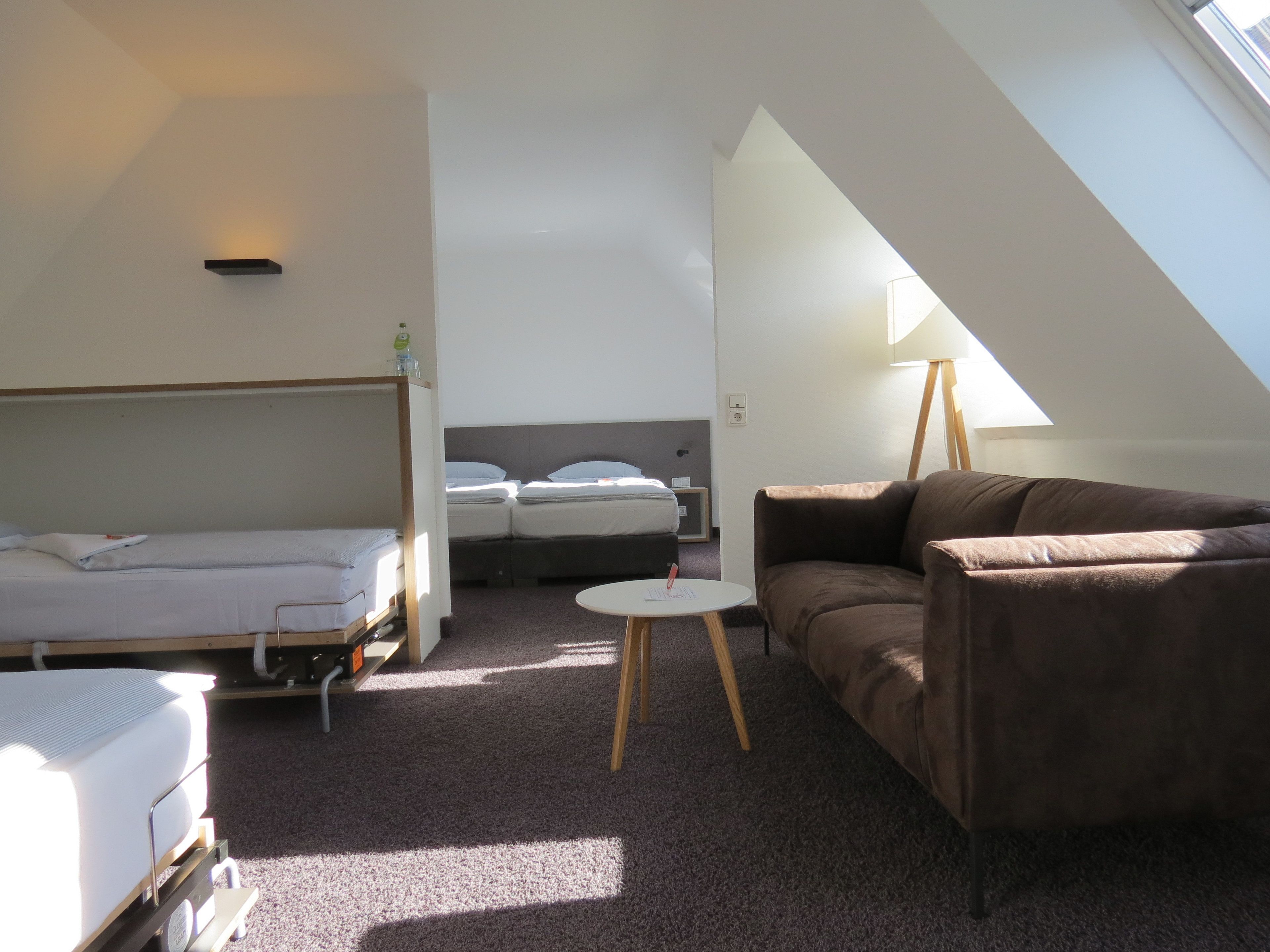 Quarto de hotel compacto com uma cama beliche, uma cama de solteiro, um sofá marrom, uma pequena mesa redonda e uma luminária.