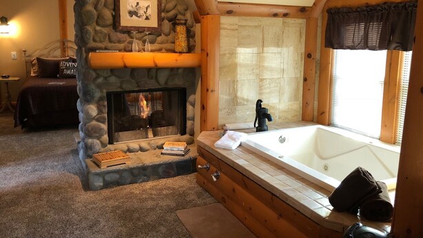 A log cabin-style room with a stone fireplace, a large bathtub, and a cozy bed with a 'Adventure Awaits' pillow. - Lazy Cloud (Lake Geneva)