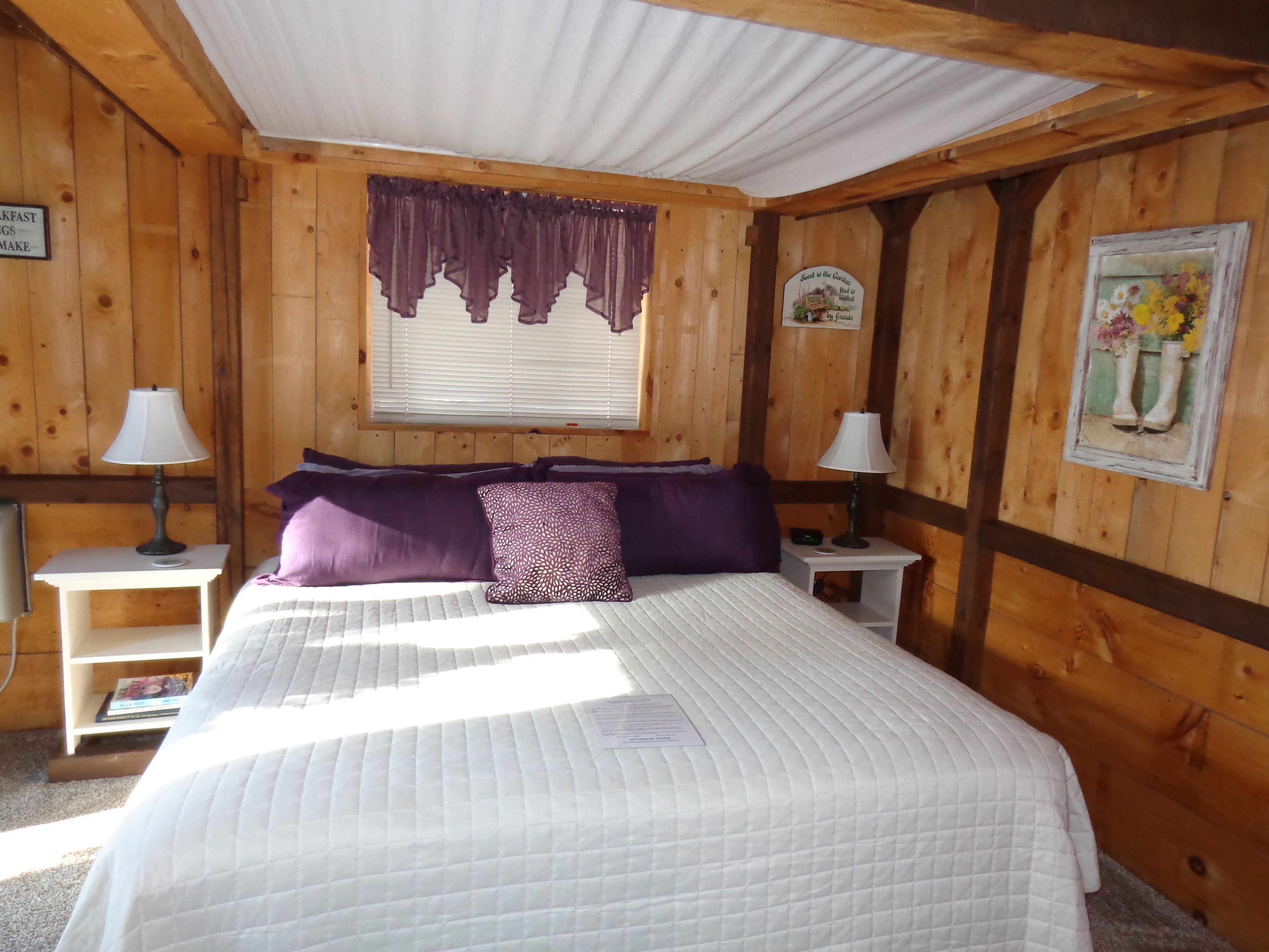 Una habitación de madera con una cama, dos mesitas de noche con lámparas, una ventana con cortinas y un cuadro enmarcado en la pared.