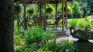 A wooden gazebo with a bench, surrounded by lush greenery and a stone planter. - Lazy Cloud (Lake Geneva)