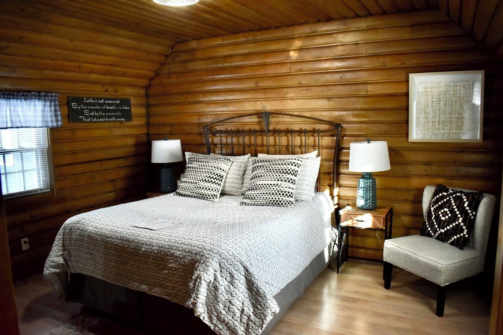 Un dormitori d'una cabana de fusta acollidora, amb un llit, dues taules de nit amb làmpades, una cadira i un cartell al mur.
