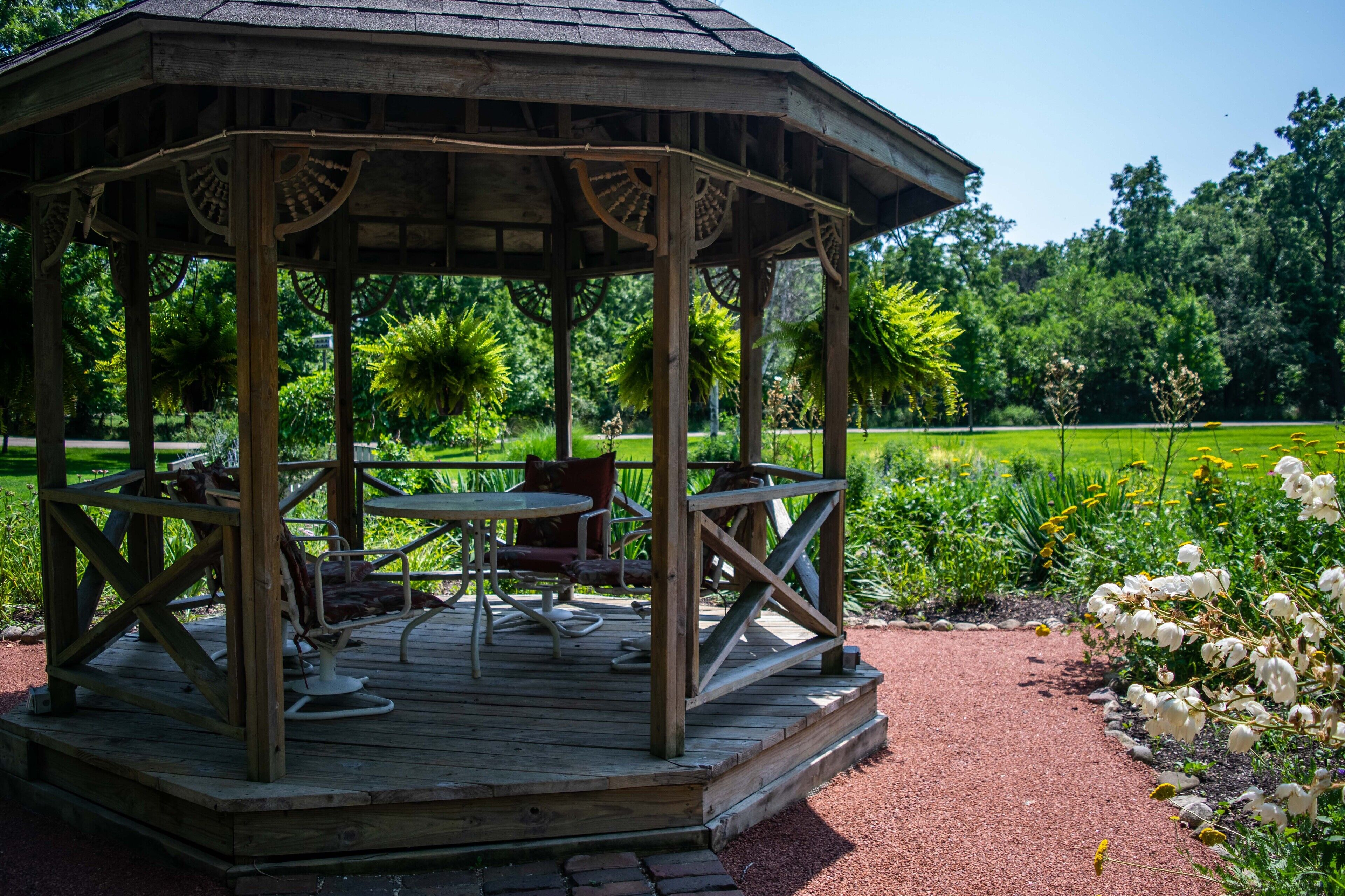 Ein hölzerner Pavillon mit Tisch und Stühlen, umgeben von einem Garten mit verschiedenen Pflanzen und Blumen.