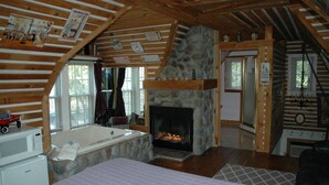 A log cabin interior with a stone fireplace, a large bed, a bathtub, and a kitchen area. - Lazy Cloud (Lake Geneva)
