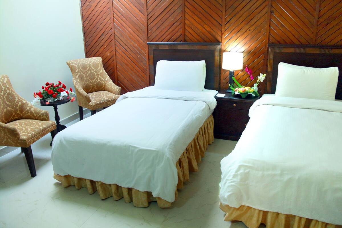 A hotel room with two beds, a wooden headboard, a nightstand with a lamp, and two armchairs.