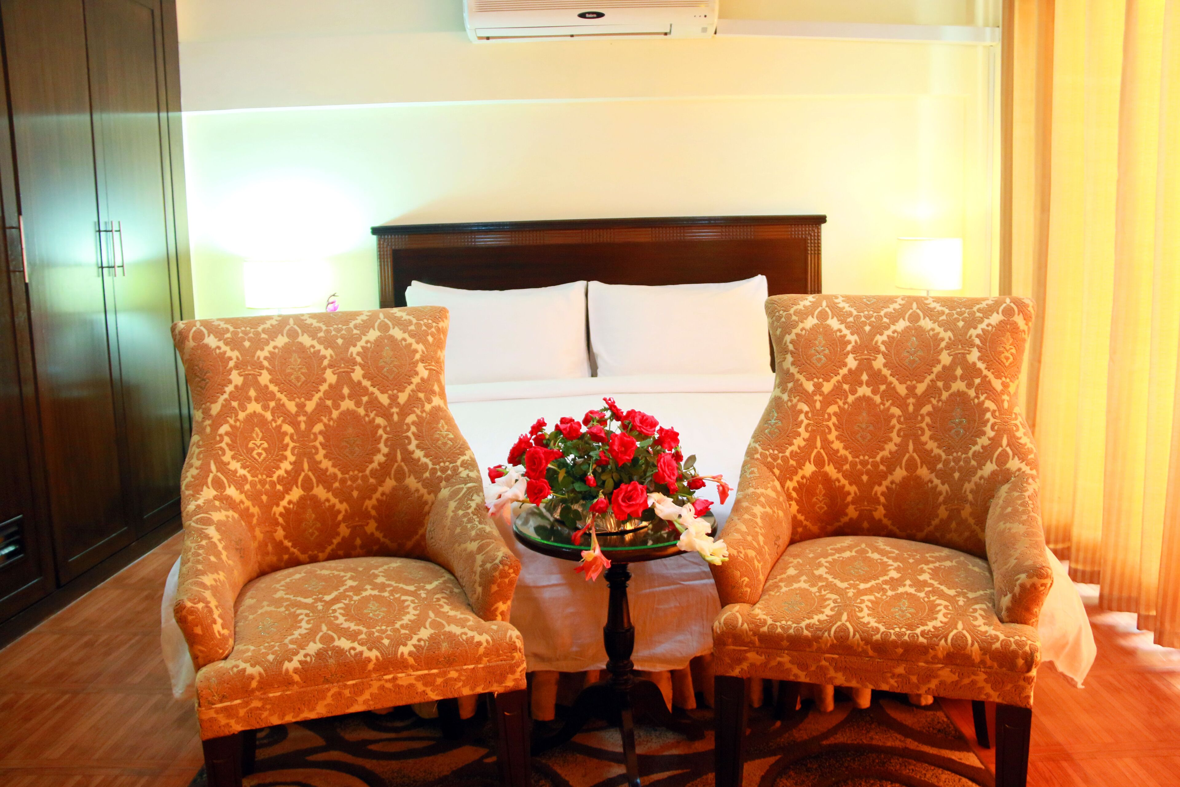 Une chambre d’hôtel avec un lit, deux fauteuils à motifs, une petite table sur laquelle est posé un vase de fleurs et un plancher en bois.